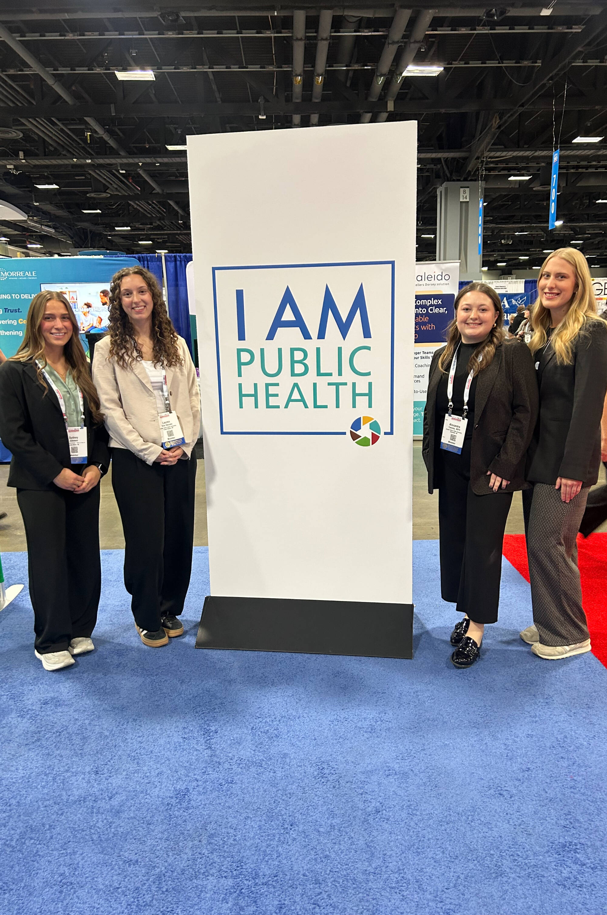 Four, smiling and professionally-dressed MPH students stand alongside a tall banner that reads, "I AM PUBLIC HEALTH" while attending a national conference.